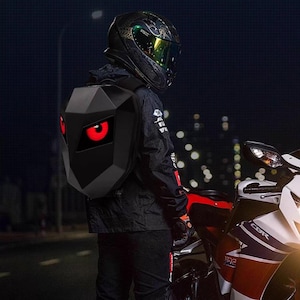 May include: Black motorcycle backpack with a geometric design and red glowing eye-shaped lights. The backpack is worn by a person wearing a black jacket and helmet, standing near a motorcycle at night.