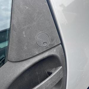 May include: A close-up of a silver car door with a black door handle and a black plastic cap with a small symbol on it.