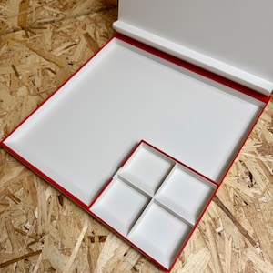 May include: A white, rectangular box with a red trim and a hinged lid. The box contains a large tray and a smaller tray divided into four square compartments. The box is open, revealing the interior.