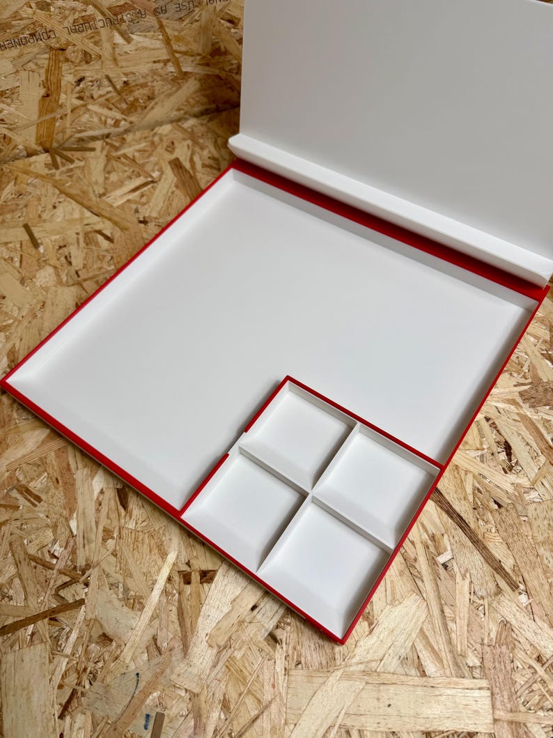 May include: A white, rectangular box with a red trim and a hinged lid. The box contains a large tray and a smaller tray divided into four square compartments. The box is open, revealing the interior.