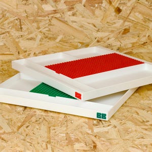 May include: Two white trays with built-in Lego baseplates. One tray has a red baseplate, the other a green one. The trays are stacked on top of each other. The trays have the letters "BB" in red and green on the corners.