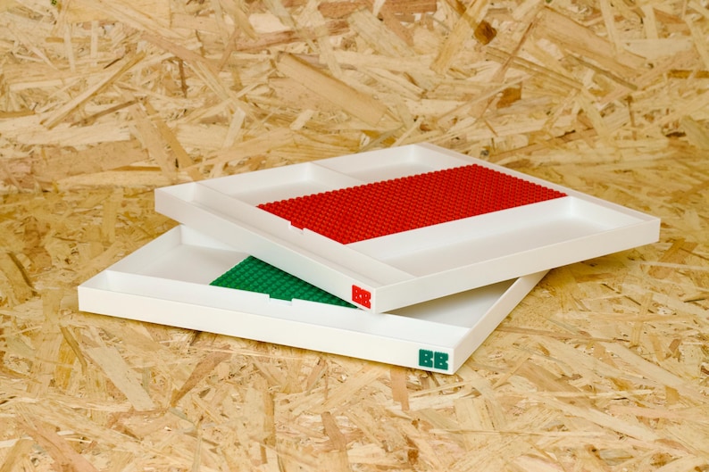 May include: Two white trays with built-in Lego baseplates. One tray has a red baseplate, the other a green one. The trays are stacked on top of each other. The trays have the letters "BB" in red and green on the corners.