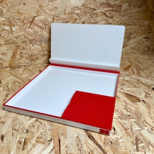 May include: An open, rectangular box with a white interior and a red border. The box has a red, textured pocket on the right side. The lid is white and open, revealing the interior. The box is on a wood-grain surface.