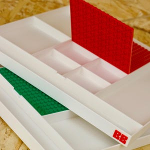 May include: White plastic storage trays with red and green building blocks. The trays have multiple compartments and are designed to hold small items. The red and green blocks are placed inside the trays. The trays have the letters "BB" in red and green.