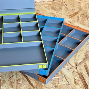 May include: Three gray rectangular storage organizers with multiple compartments. Each organizer features a distinct colored rim and matching logo: lime green, blue, and orange. The organizers are stacked diagonally, showcasing their internal grid structure.