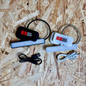 Puede incluir: Dos luces LED Brick Buddy, una negra y otra blanca, con l&aacute;mparas de cuello de cisne flexibles y cables de carga USB. La luz negra tiene el texto "BB BRICK BUDDY" en rojo y blanco. La luz blanca tiene el mismo texto.