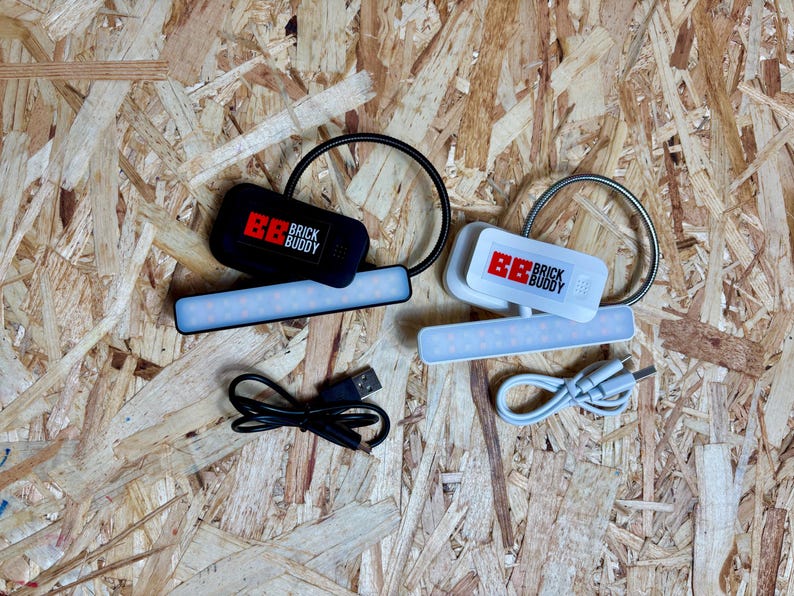 Puede incluir: Dos luces LED Brick Buddy, una negra y otra blanca, con l&aacute;mparas de cuello de cisne flexibles y cables de carga USB. La luz negra tiene el texto "BB BRICK BUDDY" en rojo y blanco. La luz blanca tiene el mismo texto.