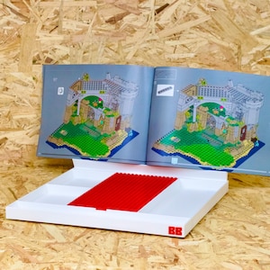 May include: Open instruction manual for a building set, displaying a detailed architectural illustration. A white tray with red building blocks sits below the manual. The background is a light brown wood.