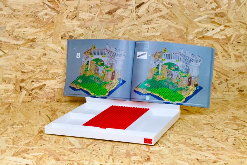 May include: Open instruction manual for a building set, displaying a detailed architectural illustration. A white tray with red building blocks sits below the manual. The background is a light brown wood.