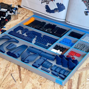 May include: A gray plastic sorting tray with multiple compartments containing various blue, black, white, gray, and red plastic building bricks and components, organized for assembly alongside an open instruction manual and a partially built model.