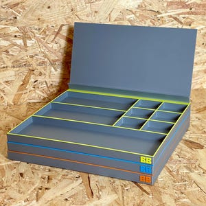 May include: A stack of three gray storage boxes with a hinged lid on the top unit. Each box features a colored rim in lime green, blue, or orange, with matching small square logos on the front corners. The interior has multiple compartments.