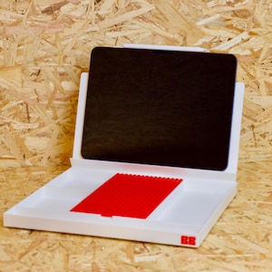 May include: A white laptop-shaped case with a black screen and a red rectangular insert. The case has a minimalist design with the letters "BB" in red on the lower right corner. The background is a light brown wood.