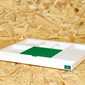 May include: A white plastic organizer tray with multiple compartments and a green base for building blocks. The tray has a rectangular shape and is placed on a wooden surface. The initials "BB" are printed in green on the lower right corner.