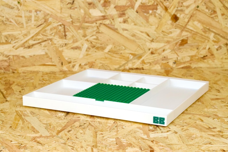 May include: A white plastic organizer tray with multiple compartments and a green base for building blocks. The tray has a rectangular shape and is placed on a wooden surface. The initials "BB" are printed in green on the lower right corner.