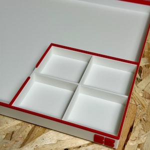 May include: A white tray with a red trim and four square compartments. The tray is on a wooden surface. The red trim is on the edges of the tray and around the compartments.