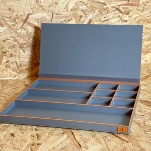 May include: A gray rectangular storage organizer with a hinged lid and multiple interior compartments. The edges of the tray and dividers are accented with bright orange trim. A small orange logo with two letters is visible on the bottom right corner.