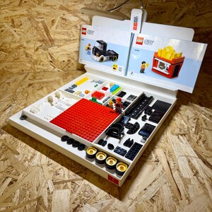 Puede incluir: Una bandeja blanca con varios ladrillos y componentes de LEGO City, incluyendo piezas rojas, negras, amarillas y blancas. Se ven dos folletos de instrucciones, uno con un cami&oacute;n negro y otro con un puesto de patatas fritas.