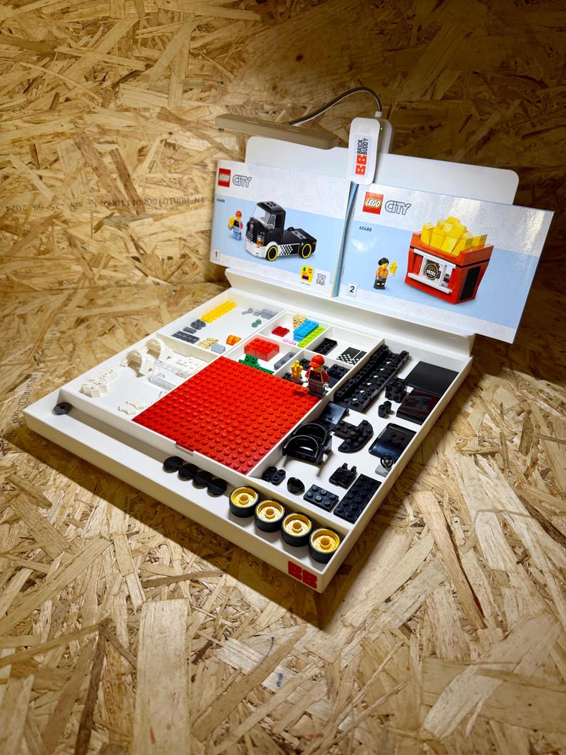 Puede incluir: Una bandeja blanca con varios ladrillos y componentes de LEGO City, incluyendo piezas rojas, negras, amarillas y blancas. Se ven dos folletos de instrucciones, uno con un cami&oacute;n negro y otro con un puesto de patatas fritas.