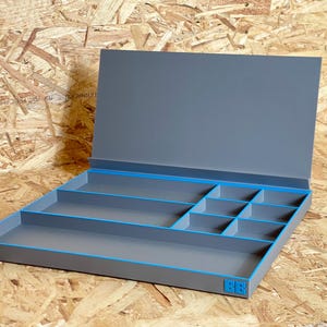 May include: A grey storage box with blue trim and compartments. The box has a hinged lid and is set against a wood-grain background. The box is designed for organising small items.