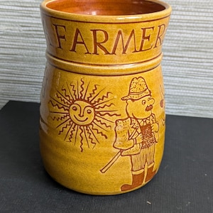 May include: A ceramic jar with a yellow-brown glaze. The word "FARMER" is printed in brown letters around the top. The jar features a smiling sun and a farmer illustration. The farmer is holding a tool and wearing a hat.