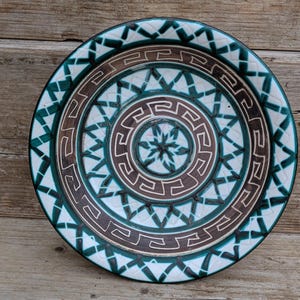 May include: A round ceramic plate featuring a central star pattern surrounded by concentric rings of brown Greek key motifs and a wide outer border of dark green and white geometric shapes arranged in a repeating crisscross pattern.