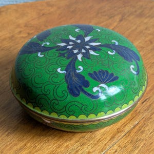 May include: A round, lidded metal box featuring a vibrant green enamel surface decorated with intricate dark blue and white floral patterns and swirling filigree details. The container has a smooth, polished metallic rim and a domed top.