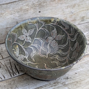 May include: A round ceramic bowl with a dark olive green glaze featuring an etched white floral and vine pattern. The interior and exterior surfaces display a smooth, glossy finish with intricate botanical line work carved into the clay.