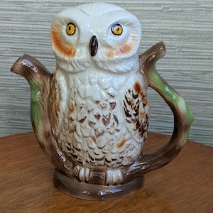May include: A ceramic owl-shaped teapot. The teapot is mainly white with brown, orange, and green details. The owl has yellow eyes, and the handle and spout are designed to resemble tree branches. The teapot is on a wooden surface.
