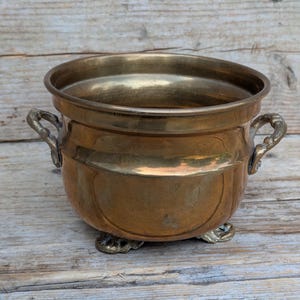 May include: A polished brass bowl with a wide circular opening, featuring two ornate side handles and three small decorative feet. The metal surface displays a warm, metallic gold tone with subtle variations in colour and a smooth, reflective finish.
