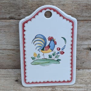 May include: A white ceramic chopping board with a scalloped red border and a central hand-painted illustration of a colourful rooster standing on a green patch of grass, featuring blue, yellow, and red plumage, with a circular hole at the top.