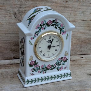 May include: A white ceramic mantel clock with a rounded top, featuring a floral and butterfly pattern in pink, green, and yellow. The clock face has black Roman numerals, a gold-tone bezel, and the Portmeirion brand name printed in black.