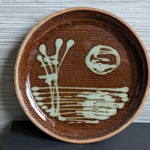 May include: A round, brown ceramic plate with a textured surface. The plate features a light green abstract design of a tree, sun, and water. The rim of the plate is a lighter brown colour.