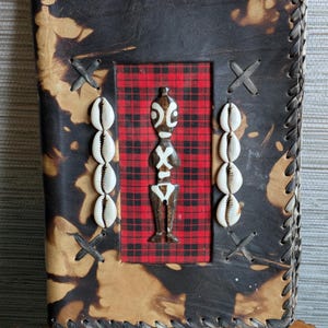 May include: A handcrafted leather journal with a black and tan spotted cover. The journal features a red plaid insert with a wooden figurine and cowrie shells. The edges are stitched with a decorative pattern.
