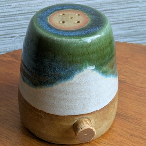 May include: A handmade ceramic salt shaker with a cork stopper. The shaker is a mix of colours, including green, blue, white, and brown. The top has several holes for dispensing salt.