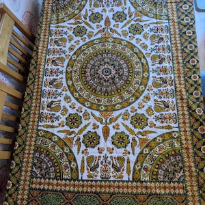 May include: A rectangular white tablecloth with a detailed pattern in gold, green, and brown. The design includes floral and geometric shapes, such as circular medallions and border patterns. The tablecloth is likely made of fabric, suitable for a table.