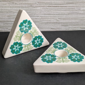 May include: Two triangular, off-white candle holders. Each has a circular opening for a candle and is decorated with teal floral and leaf patterns. The candle holders are on a dark surface.