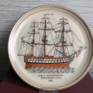 May include: Round ceramic plate with a beige speckled background and a brown rim. The plate features a detailed illustration of a tall ship with brown masts and sails, and the text "H.M.S. AGA MEMNON Built at Buckler's Hard 1781".