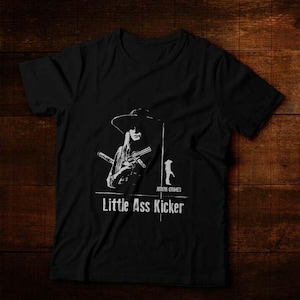 Może przedstawiać: Czarny t-shirt z białą grafiką kobiety w kapeluszu trzymającej dwa pistolety, z napisem "Little Ass Kicker" poniżej. Koszulka zawiera również napis "Judith Grimes" i małą ilustrację.