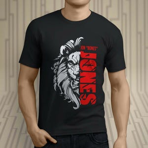 Puede incluir: Camiseta negra con un gráfico de león y el nombre "JONES" en texto vertical rojo. La cara del león está sombreada a la mitad en blanco y gris, con el texto "JON "BONES" encima de la cabeza del león. La camiseta tiene cuello redondo.