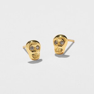 May include: A pair of gold-colored skull stud earrings. Each skull has two clear, round gemstones for eyes and a detailed jawline. The earrings are set against a white background.