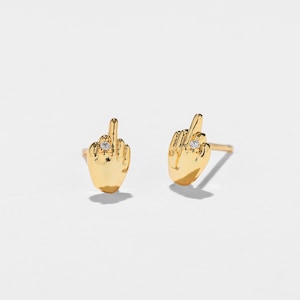 May include: A pair of gold-coloured stud earrings in the shape of a hand with the middle finger extended. Each earring has a small, clear gemstone. The earrings are set against a white backdrop.
