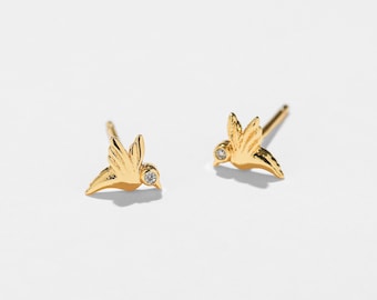 Pode incluir: Um par de brincos de pressão dourados em forma de beija-flor. Cada brinco apresenta um design detalhado de beija-flor com um pequeno detalhe de pedra preciosa transparente. Os brincos estão sobre um fundo branco.
