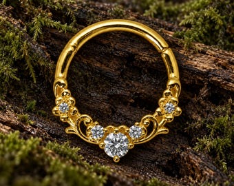 Pode incluir: Um anel de aro dourado com um segmento curvo decorativo adornado com cinco pedras preciosas transparentes de corte redondo. O metal apresenta padrões florais e de volutas intrincados em torno das pedras, com uma pedra central maior.