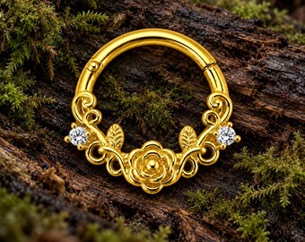 Pode incluir: Um anel circular de tom dourado com um design floral ornamentado na base. A peça central é uma rosa esculpida ladeada por duas pequenas folhas e duas pedras transparentes de corte redondo cravadas nos detalhes metálicos decorativos.