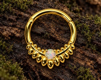Pode incluir: Um anel circular de tom dourado com um segmento decorativo adornado com uma série de pequenas contas metálicas arredondadas e uma única pedra de opala iridescente central. A peça de joalharia tem um mecanismo de fecho articulado visível.