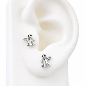 Puede incluir: Un par de pendientes de plata con forma de fantasma. Cada pendiente presenta un pequeño fantasma estilizado con los brazos extendidos y ojos negros. Los pendientes se muestran en un modelo de oreja blanco, mostrando su diseño y tamaño.