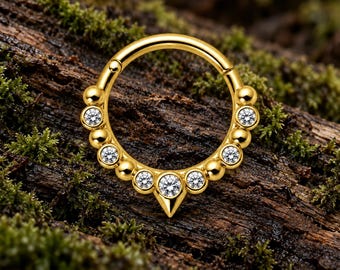 Pode incluir: Um anel circular de tom dourado com um segmento articulado e um arco inferior decorativo. O design inclui uma alternância de esferas douradas polidas e pedras transparentes de corte redondo, terminando numa ponta dourada central.
