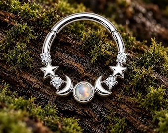 Pode incluir: Um anel circular de tom prateado com um cabochão central de opala iridescente, ladeado por duas luas crescentes prateadas, duas pequenas estrelas prateadas e quatro pedras redondas facetadas transparentes dispostas simetricamente.
