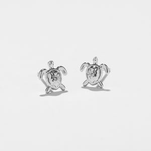 May include: A pair of silver turtle stud earrings. Each earring is a detailed miniature turtle with a small, clear gem at the shell's centre. The earrings are displayed on a white background, highlighting their design.