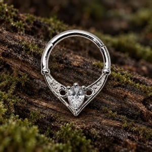 Puede incluir: Un anillo de segmento articulado en tono plateado con un adorno en forma de V. El centro de la pieza en V contiene una piedra preciosa transparente de corte en forma de pera, flanqueada por dos peque&ntilde;os huecos circulares y detalles.
